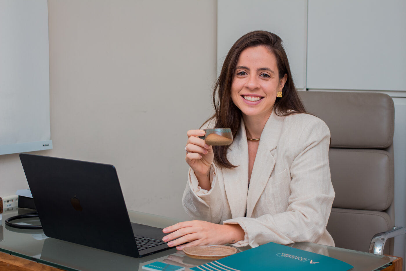 Dra. Amanda Saavedra à mesa do consultório, com o notebook aberto e uma xícara na mão
