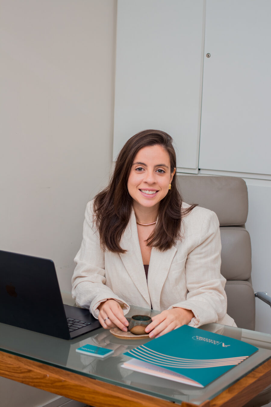 Dra. Amanda Saavedra, de blazer claro, sorrindo à mesa do consultório